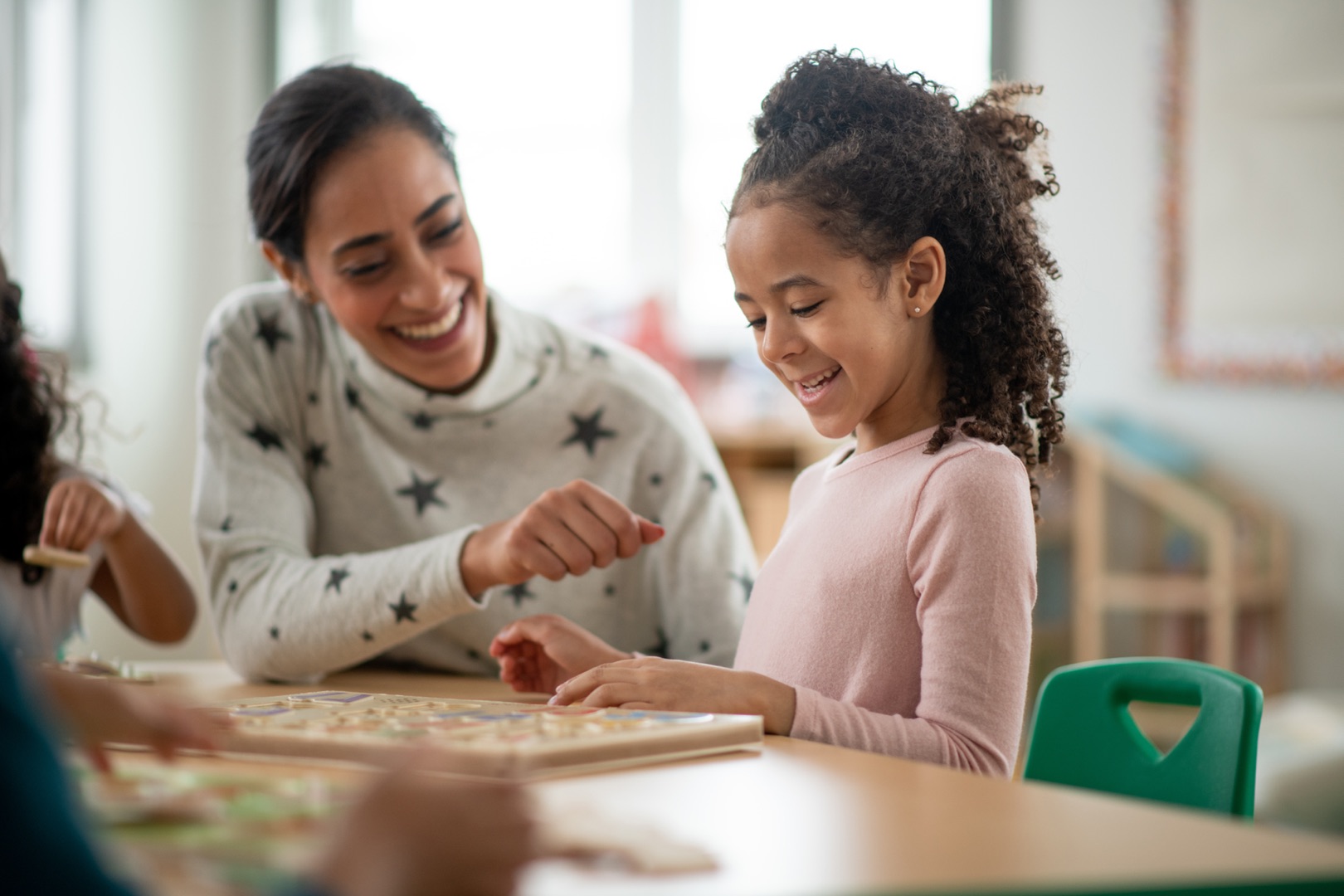 A young female daycare teacher of Indian decent, sits beside a sweet, curly-hired young girl as they look at the completed puzzle they cooperatively finished. They are each dressed casually, smiling and laughing as they sit back and enjoy looking at their finished work.